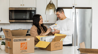 A male and female in a kitchen with moving boxes and unboxing a care package