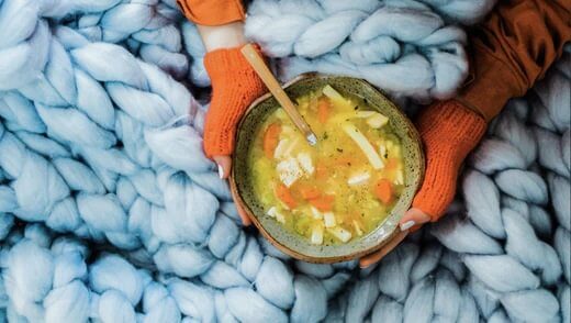 Image of two gloved hands holding a bowl of soup on a thick knit blanket.