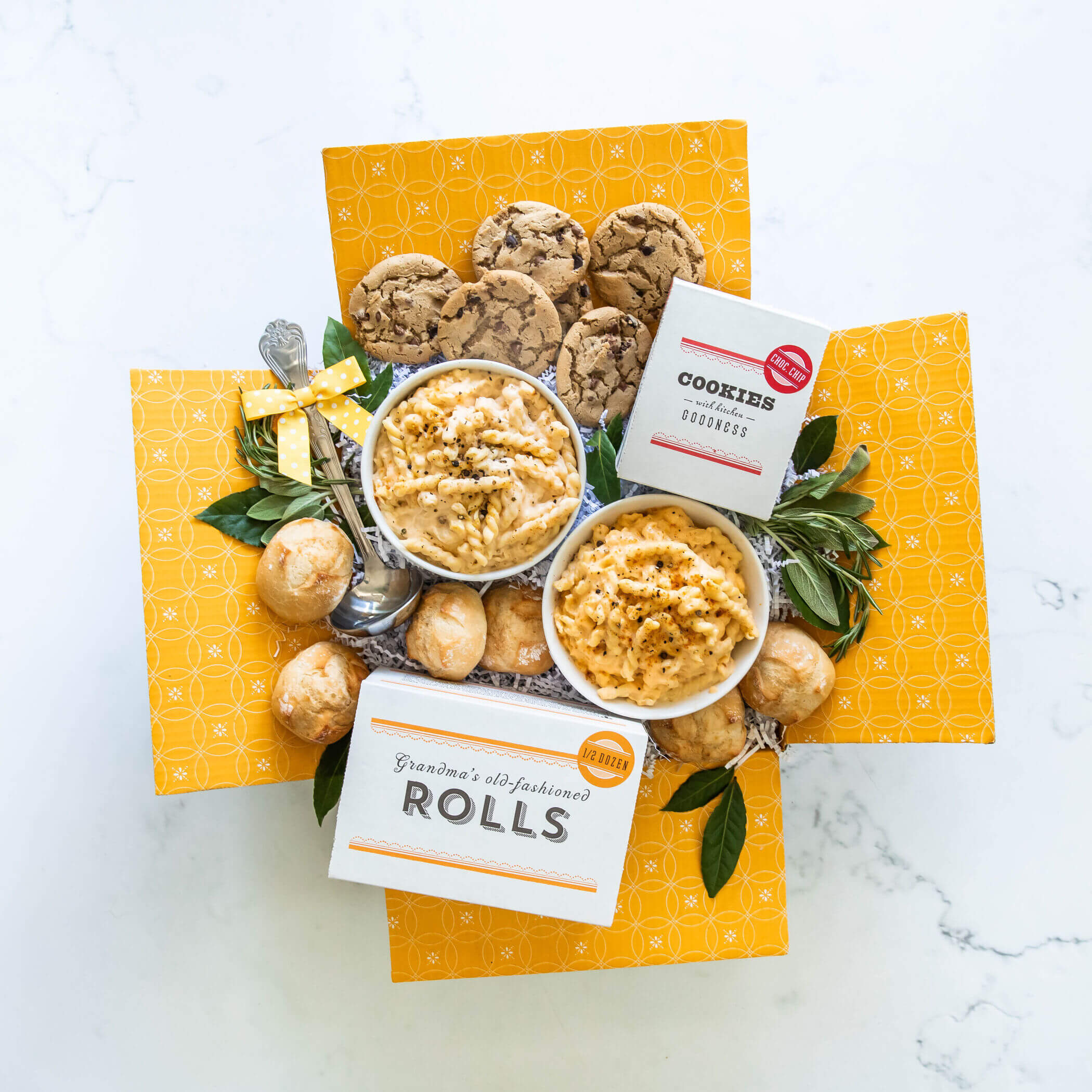Open Mac n' Cheese gift basket with two bowls of macaroni surrounded by rolls and cookies and a ladle. 