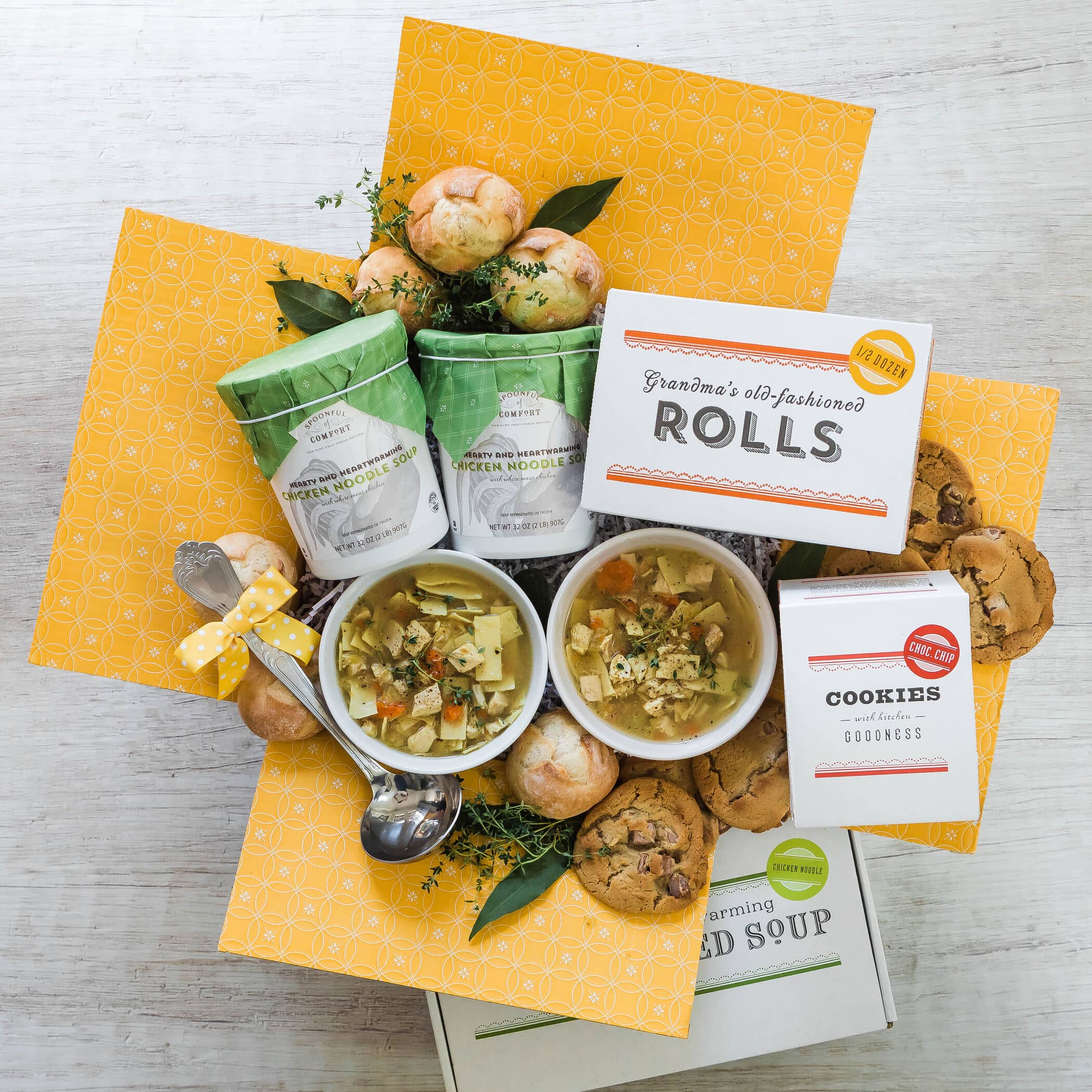 Open chicken noodle soup care package with ladle, cookies, and rolls.